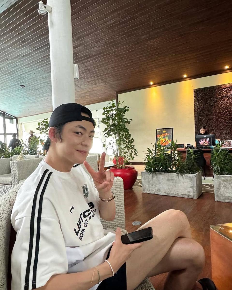 Seorang pria mengenakan kaus putih bergaris hitam dan topi hitam terbalik duduk santai di ruang lounge dengan tanaman hias di sekitarnya.