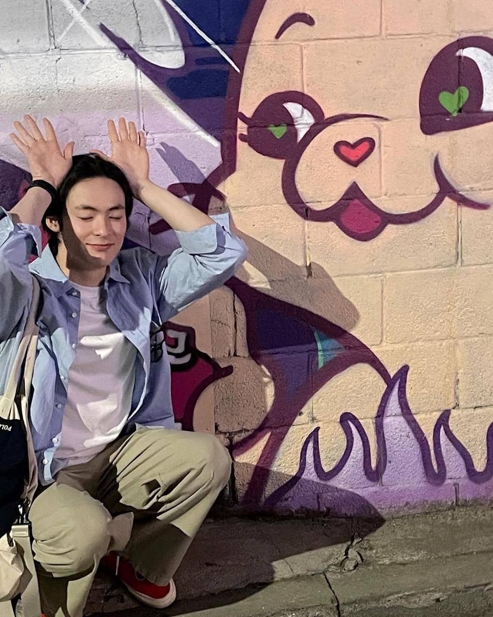Seorang pria berpose lucu dengan tangan di atas kepala di depan mural berwarna cerah bergambar karakter kartun di dinding bata.