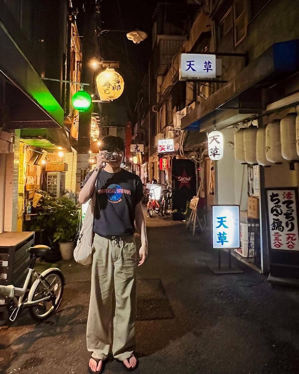 Seorang pria mengenakan kaus hitam bertulisan Universal Studios dan celana panjang krem berdiri di gang sempit dengan lampu neon Jepang pada malam hari.