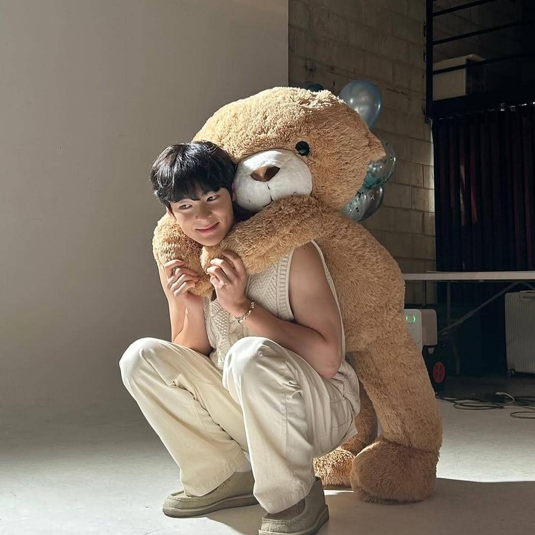 Seorang pria mengenakan outfit kasual berwarna krem sedang berpose sambil dipeluk boneka beruang besar di ruangan dengan pencahayaan lembut.