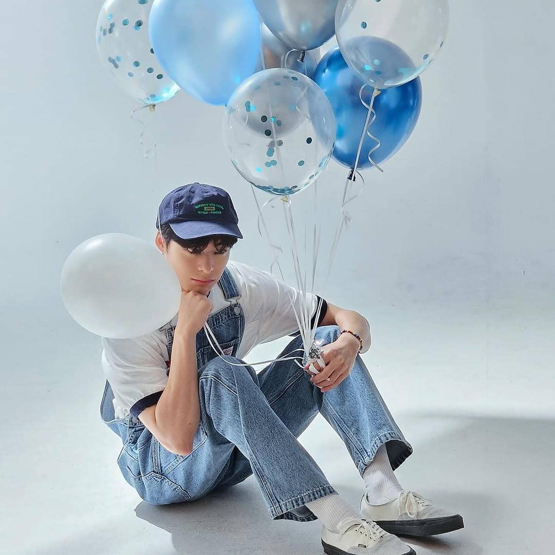 Seorang pria mengenakan kaus putih, celana overall denim, dan topi biru duduk di lantai sambil memegang balon berwarna biru dan perak.