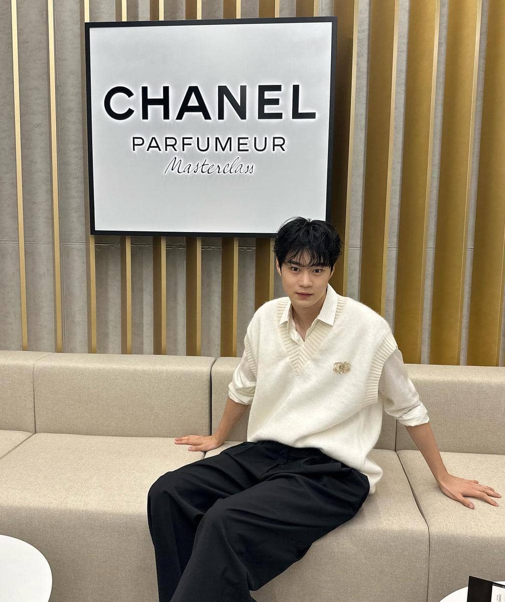 Seorang pria mengenakan kemeja putih dan rompi rajut krem duduk di sofa di depan papan bertuliskan Chanel Parfumeur Masterclass.