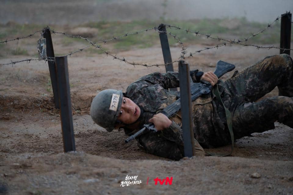 Seorang tentara berpakaian loreng merayap di bawah kawat berduri sambil memegang senapan dalam adegan drama The Legend of Kitchen Soldier.