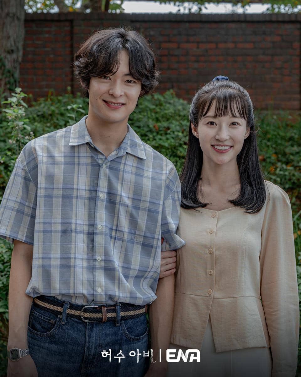 Dua pemeran drama The Scarecrow berdiri berdekatan sambil tersenyum di luar ruangan dengan latar taman dan tembok bata.