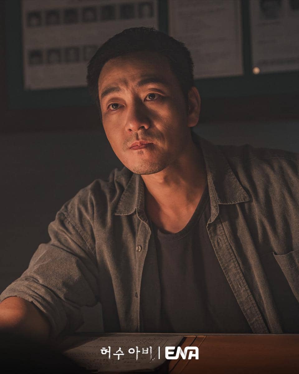 Seorang pria duduk di meja dengan pencahayaan redup, mengenakan kemeja abu-abu dan menatap serius dalam adegan drama The Scarecrow.