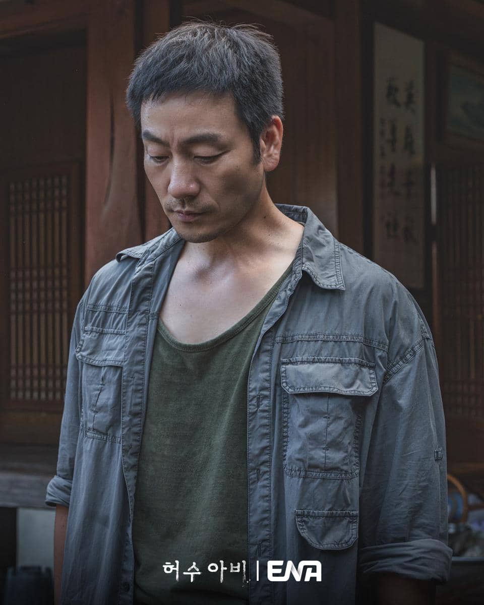 Seorang pria berwajah serius mengenakan jaket abu-abu dan kaus hijau berdiri di depan bangunan kayu tradisional Korea.