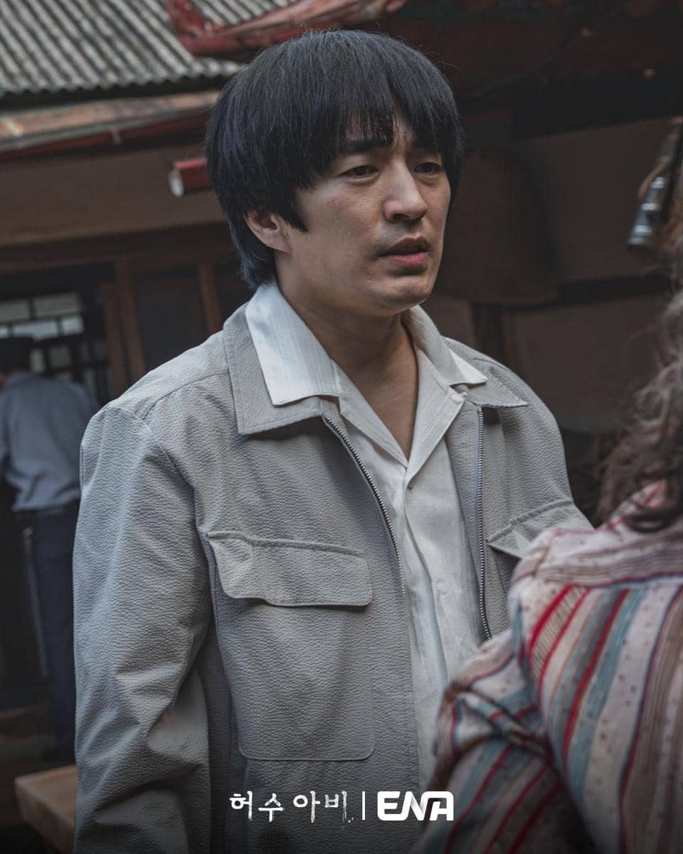 Seorang pria berambut hitam mengenakan jaket abu-abu dan kemeja putih berdiri di luar bangunan tradisional dalam adegan drama The Scarecrow.