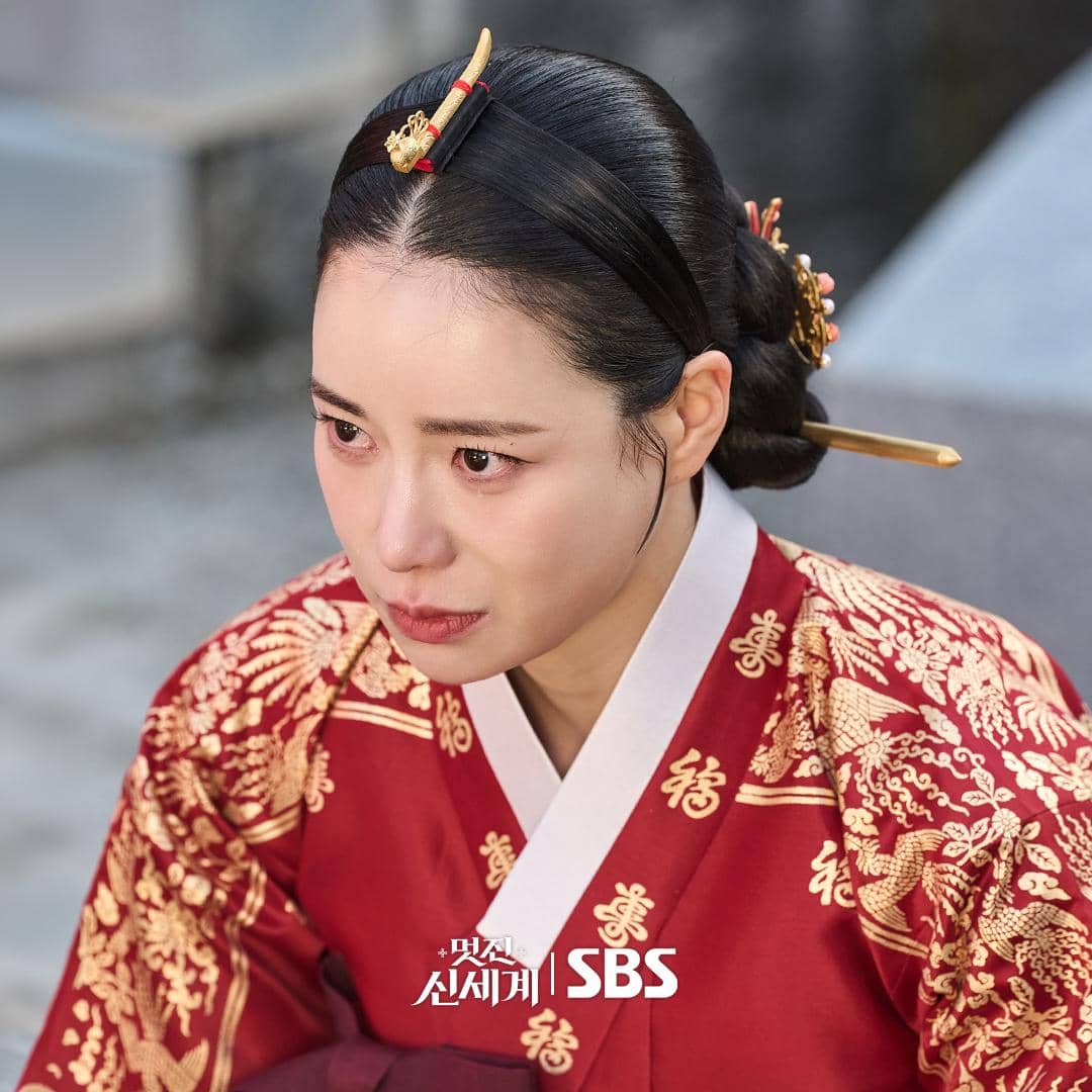 Aktris mengenakan hanbok merah berhias bordir emas dengan gaya rambut tradisional dan hiasan kepala di drama My Royal Nemesis.