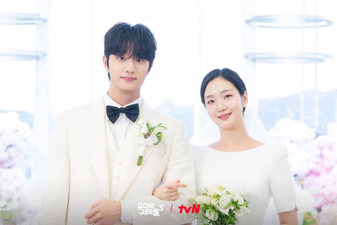 Dua pemeran utama mengenakan busana pengantin putih berdiri bersama di lokasi pernikahan dalam adegan drama Yumi's Cells 3.