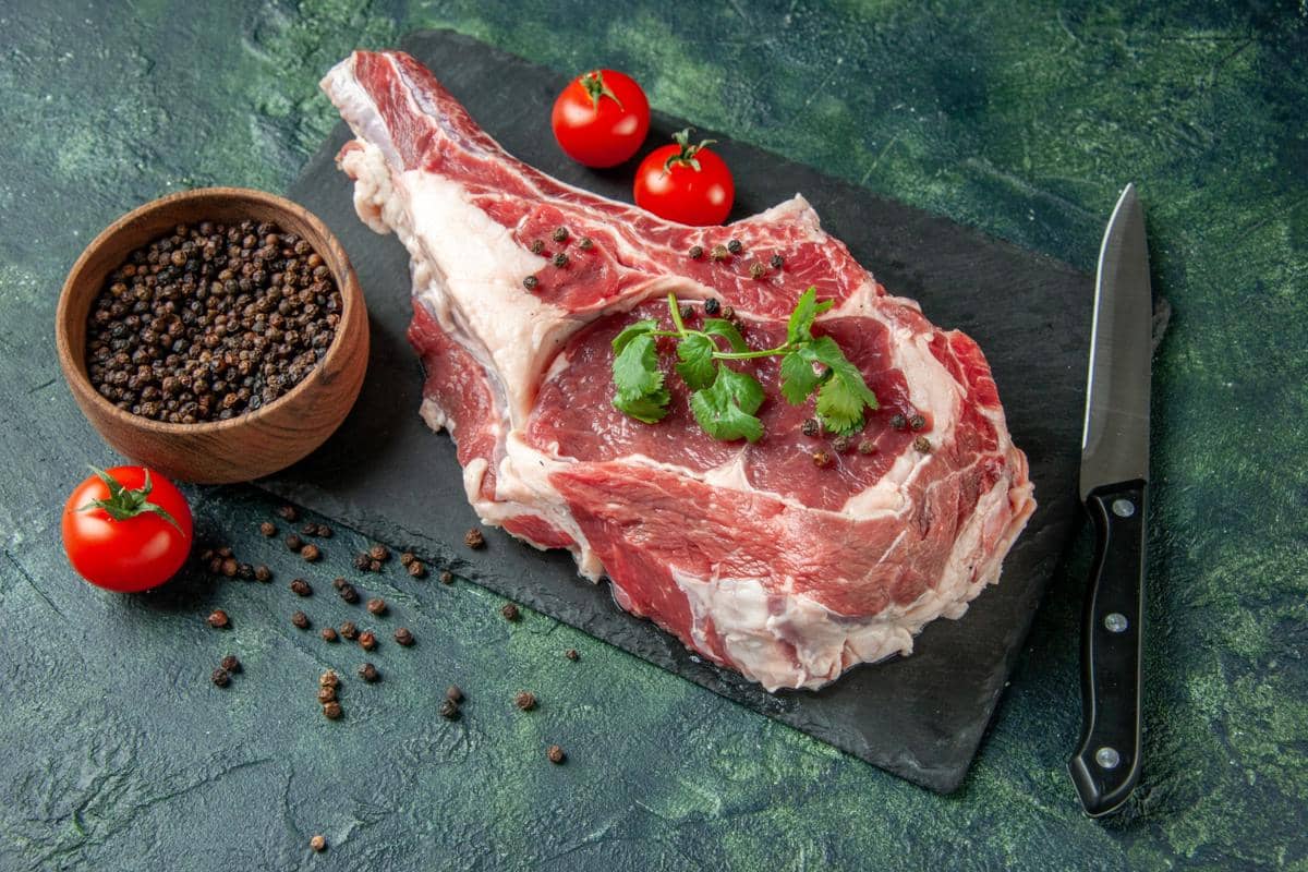 Potongan daging sapi mentah di atas papan hitam dengan taburan lada hitam, daun ketumbar, tomat ceri, dan pisau di sampingnya.