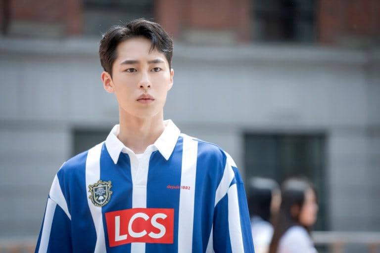 Seorang pria mengenakan seragam bergaris biru putih dengan logo LCS berdiri di luar gedung sekolah dalam adegan drama Extraordinary You.