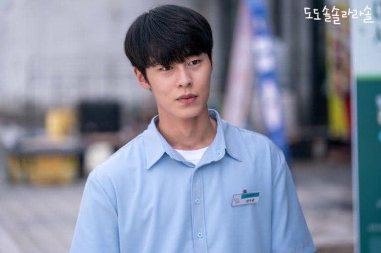 Seorang pria mengenakan seragam biru muda dengan name tag berdiri di luar ruangan dalam adegan drama Do Do Sol Sol La La Sol.