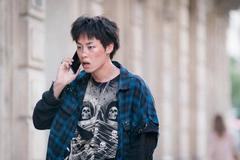 Seorang pria muda dengan wajah terluka mengenakan kemeja flanel biru dan kaus bergambar tengkorak sedang berbicara di telepon di luar ruangan.