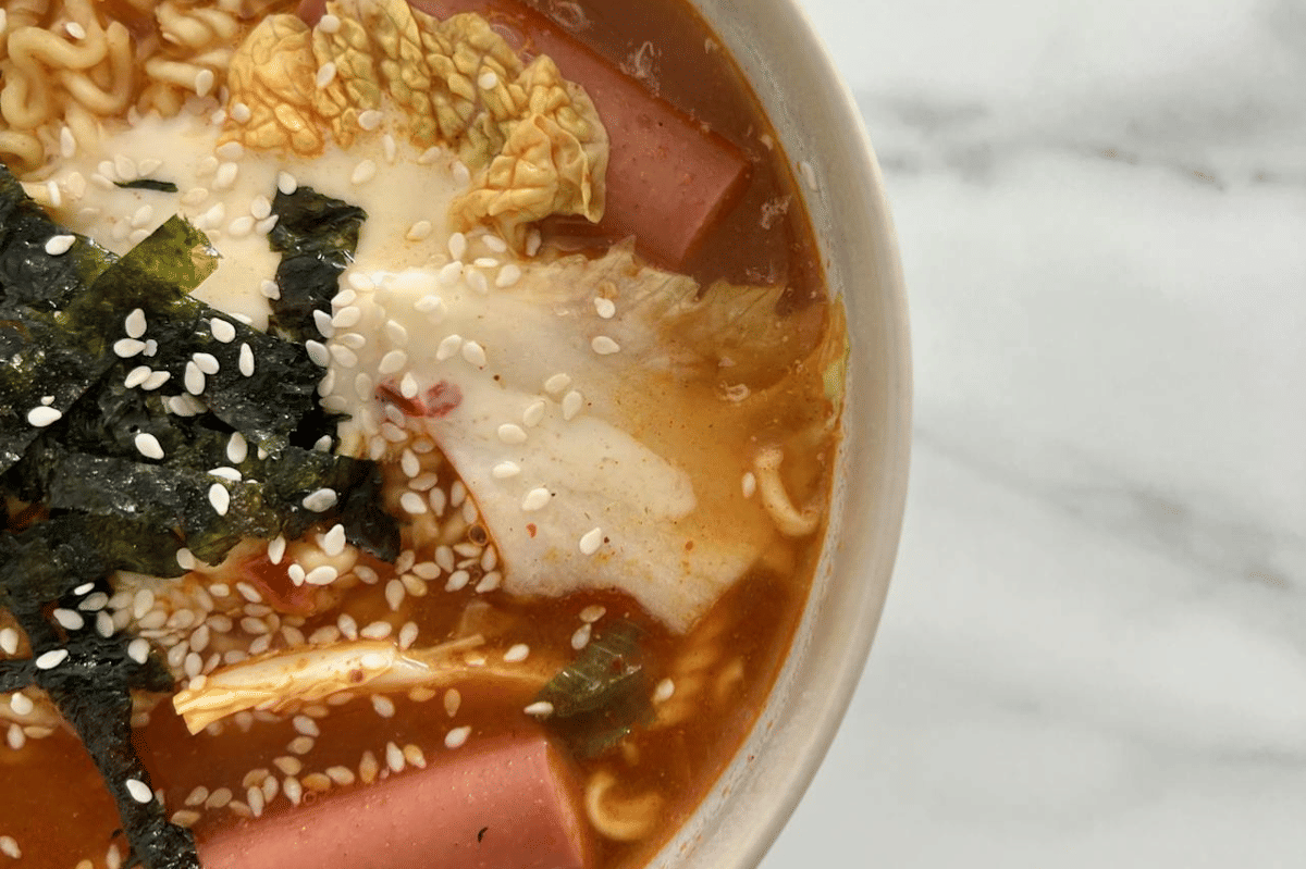 Semangkuk sausage ramen carbonara dengan kuah kental, potongan sosis, rumput laut, biji wijen, dan sayuran di atas meja marmer.