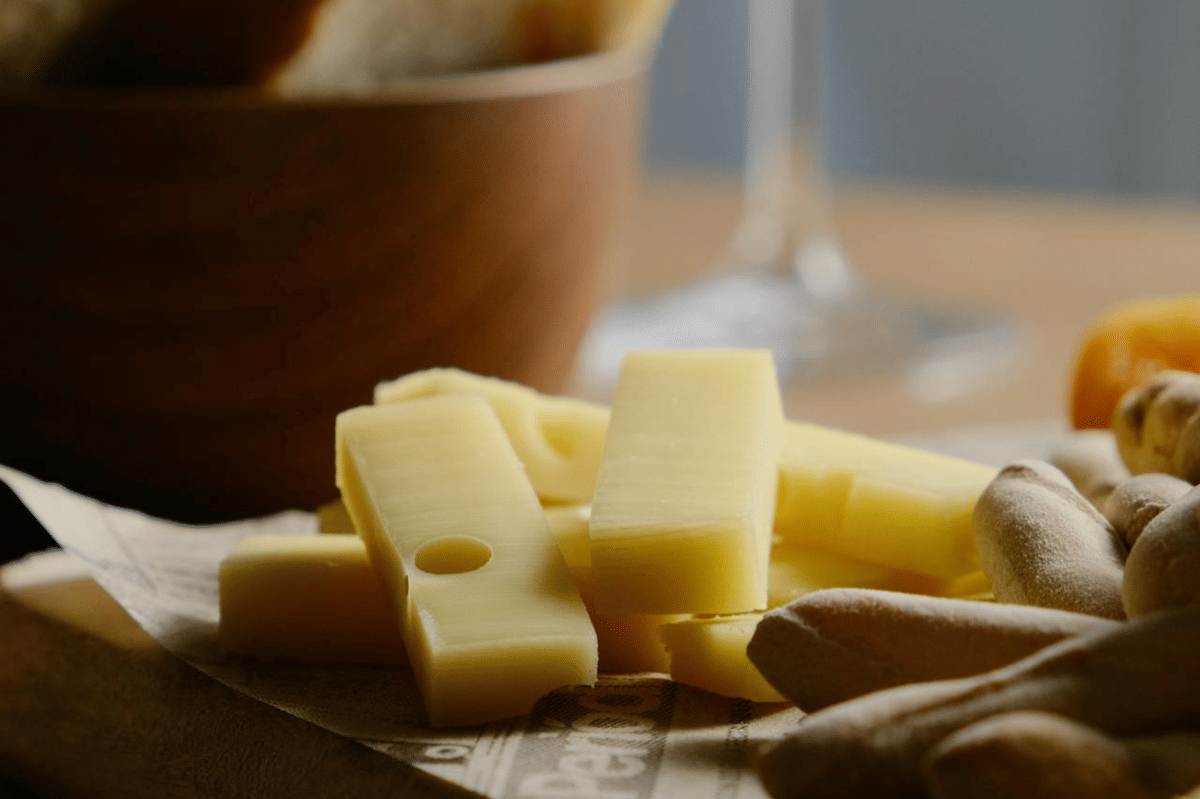 Potongan keju cheddar berwarna kuning muda tersusun di atas kertas dengan latar belakang roti dan mangkuk kayu.