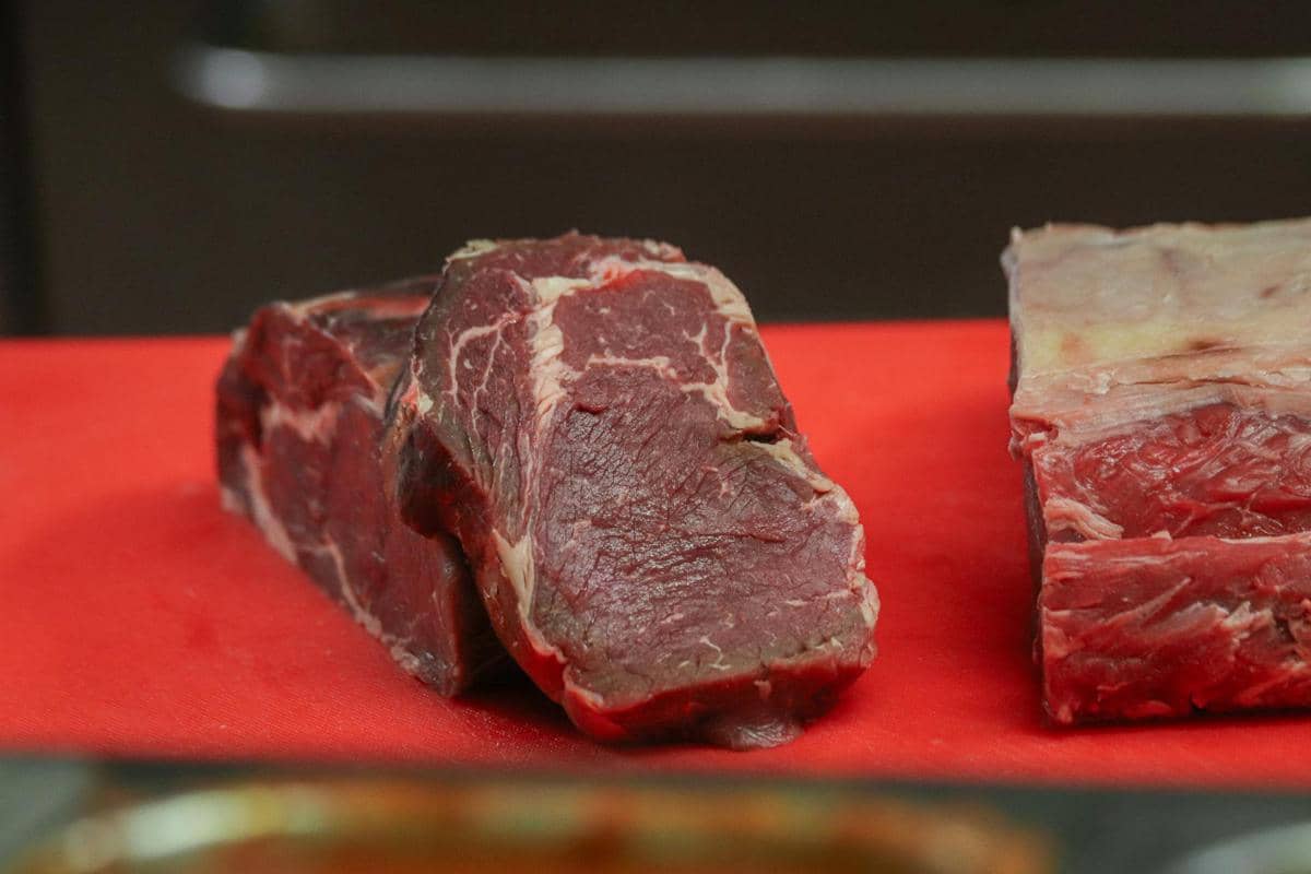 Potongan daging sapi mentah berwarna merah tua dengan serat lemak putih di atas alas berwarna merah di dapur.