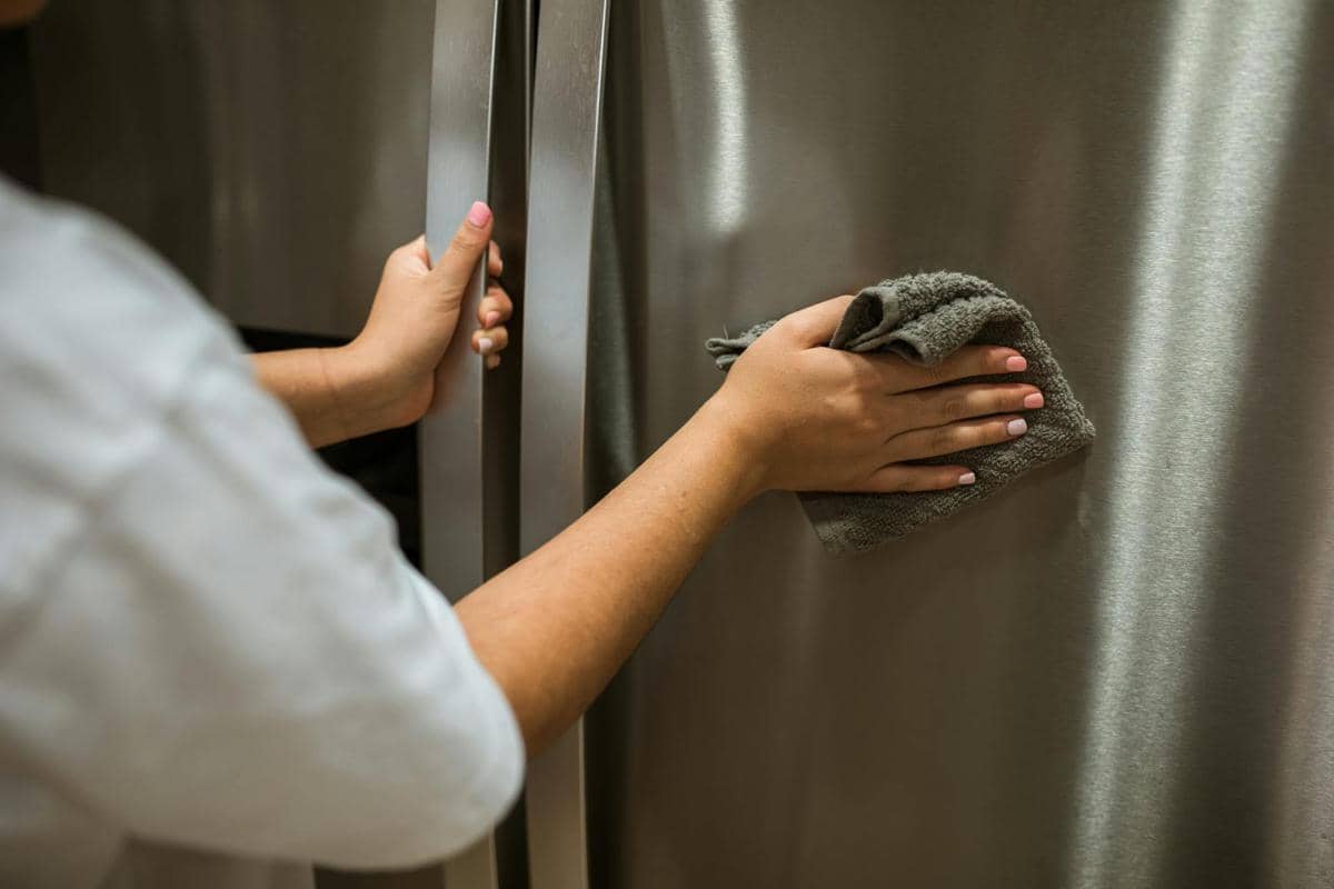 Seseorang membersihkan pintu kulkas stainless steel dengan kain lap berwarna abu-abu untuk menjaga kebersihan permukaannya.