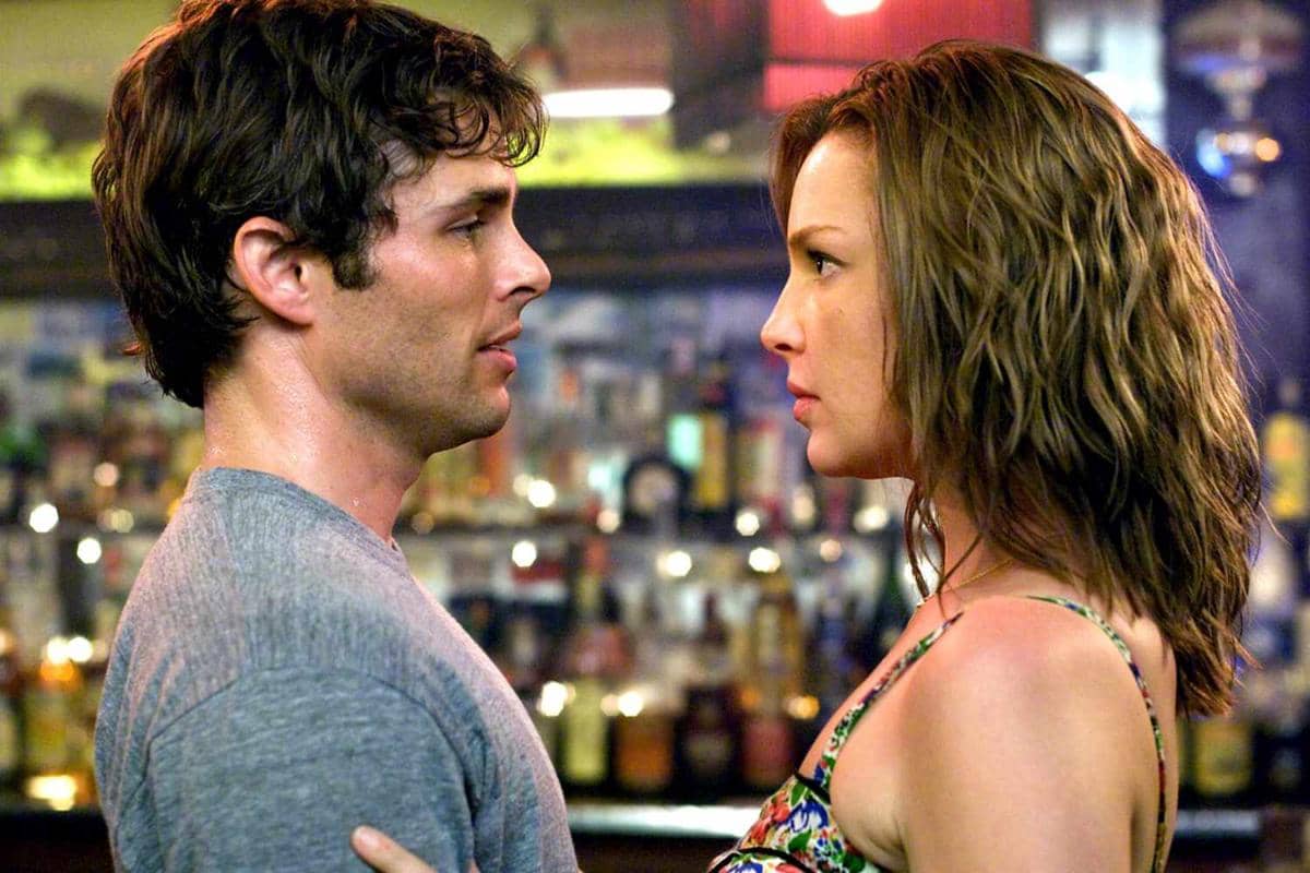 Dua karakter dalam film 27 Dresses saling menatap dengan ekspresi serius di sebuah bar yang remang dengan latar botol minuman.