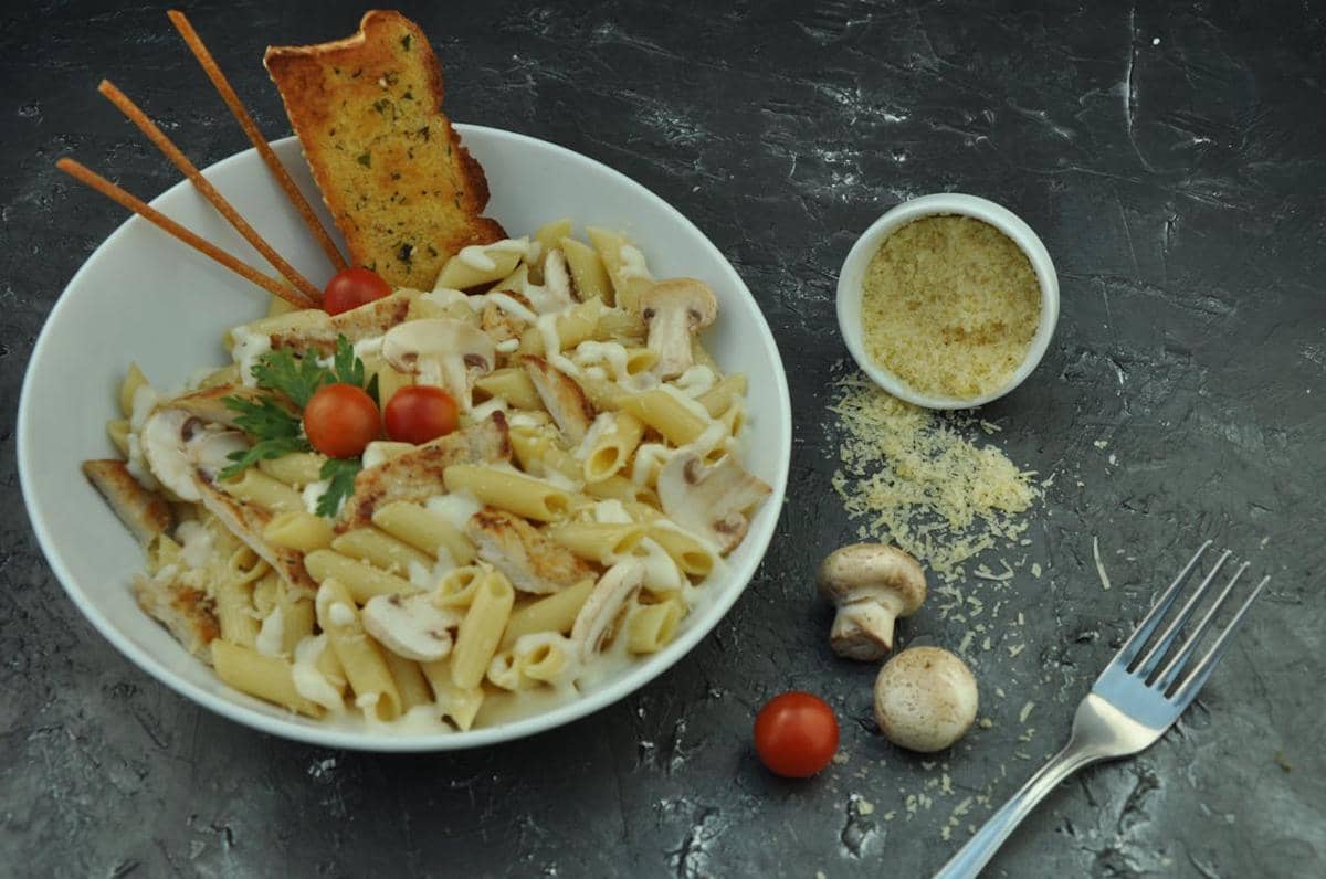 Semangkuk pasta penne dengan jamur, tomat ceri, roti panggang, dan taburan keju parmesan di atas meja gelap.