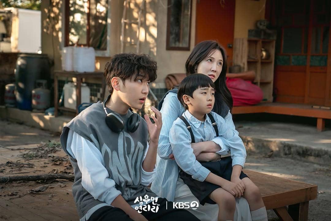 Tiga karakter duduk di halaman rumah tradisional, seorang wanita memeluk anak kecil sementara pria muda dengan headphone duduk di samping mereka.