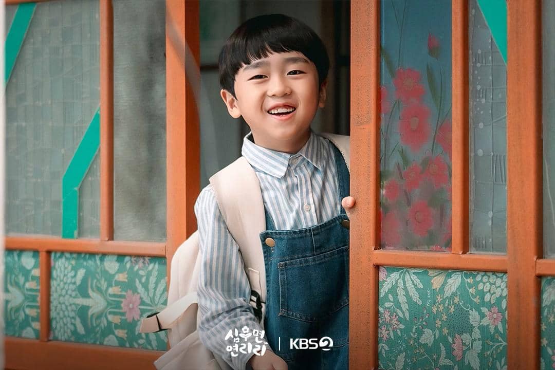 Anak laki-laki mengenakan kemeja bergaris biru dan overall denim tersenyum ceria sambil berdiri di pintu rumah dengan kaca bermotif bunga.