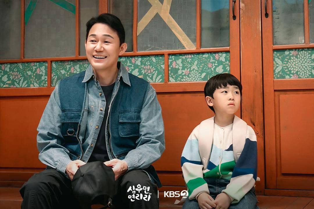Seorang pria dewasa tersenyum duduk di samping anak laki-laki kecil di depan pintu kayu dengan latar dinding bermotif bunga.