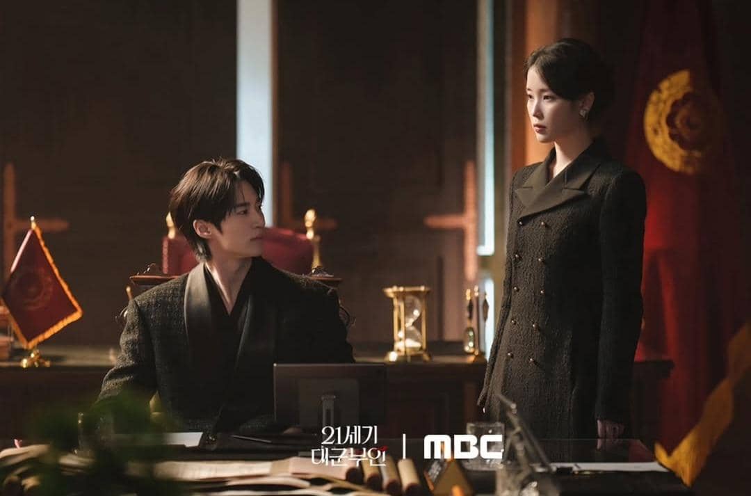 Dua karakter dalam drama Korea Perfect Crown berada di ruangan bergaya klasik, satu duduk di meja kerja dan satu berdiri mengenakan setelan hitam.