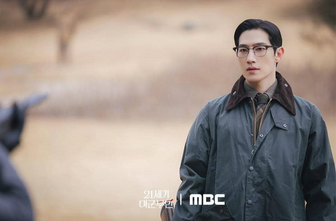 Seorang pria berjas hijau dan berkacamata berdiri di luar ruangan dengan latar padang rumput cokelat dalam cuplikan drama Perfect Crown.