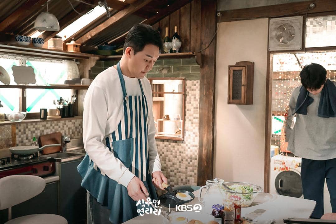 Seorang pria mengenakan celemek bergaris sedang memasak telur di dapur bergaya rustic dengan pencahayaan hangat, sementara pria lain berdiri di belakangnya.