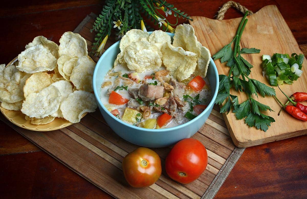 Semangkuk sop kambing bersantan dengan potongan daging, tomat, dan kerupuk di atas meja kayu bersama bahan pelengkap segar.