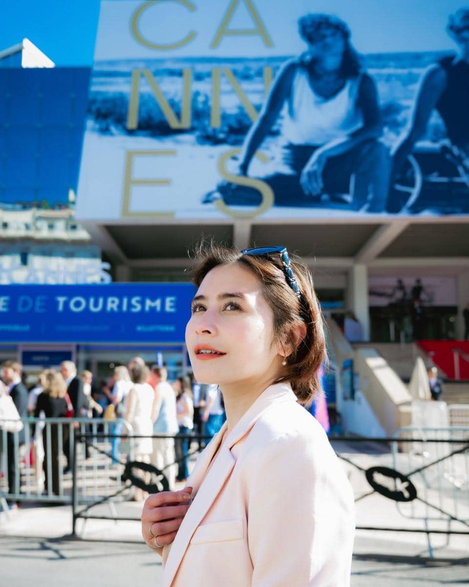 Seorang perempuan mengenakan blazer berwarna terang berdiri di area luar gedung dengan latar spanduk besar bertuliskan Cannes.