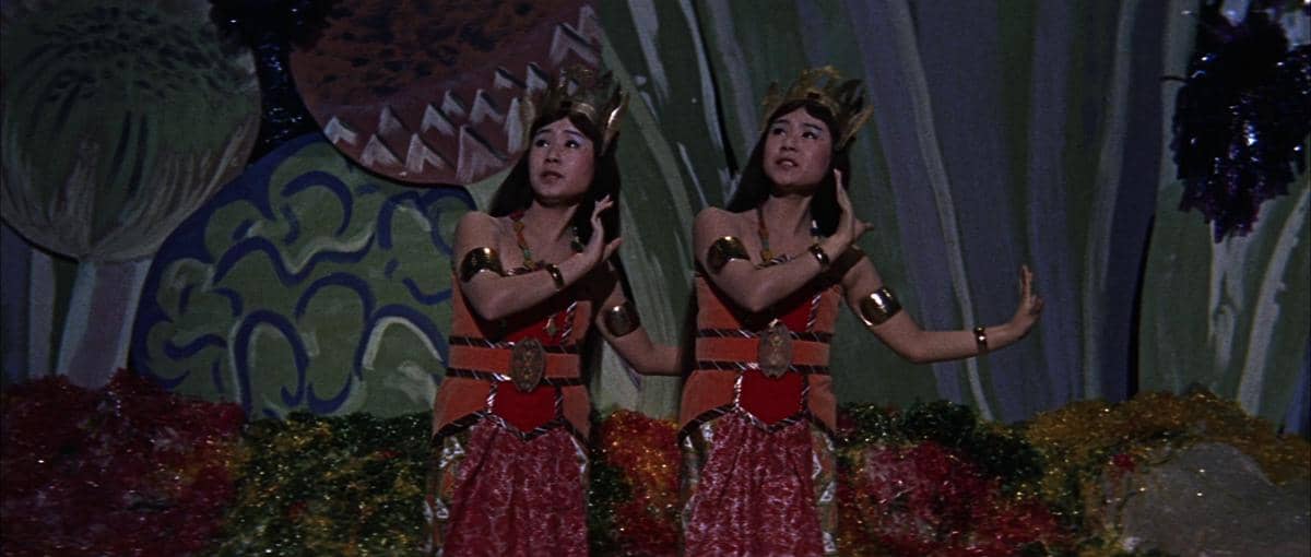 Dua peri kembar Shobijin mengenakan kostum tradisional berwarna merah dan emas, berdiri di latar berwarna cerah dengan ornamen alam.