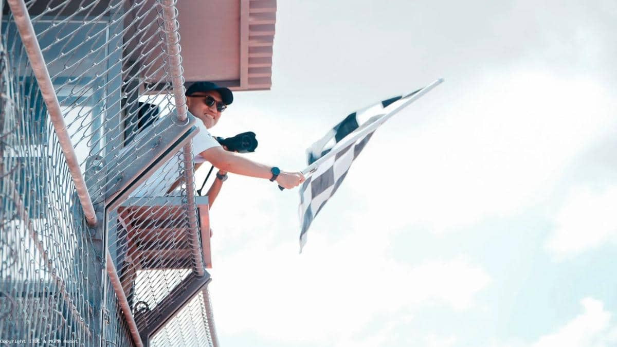 Seorang pria mengenakan kacamata hitam melambaikan bendera kotak-kotak dari menara pengawas di lintasan balap di bawah langit cerah.