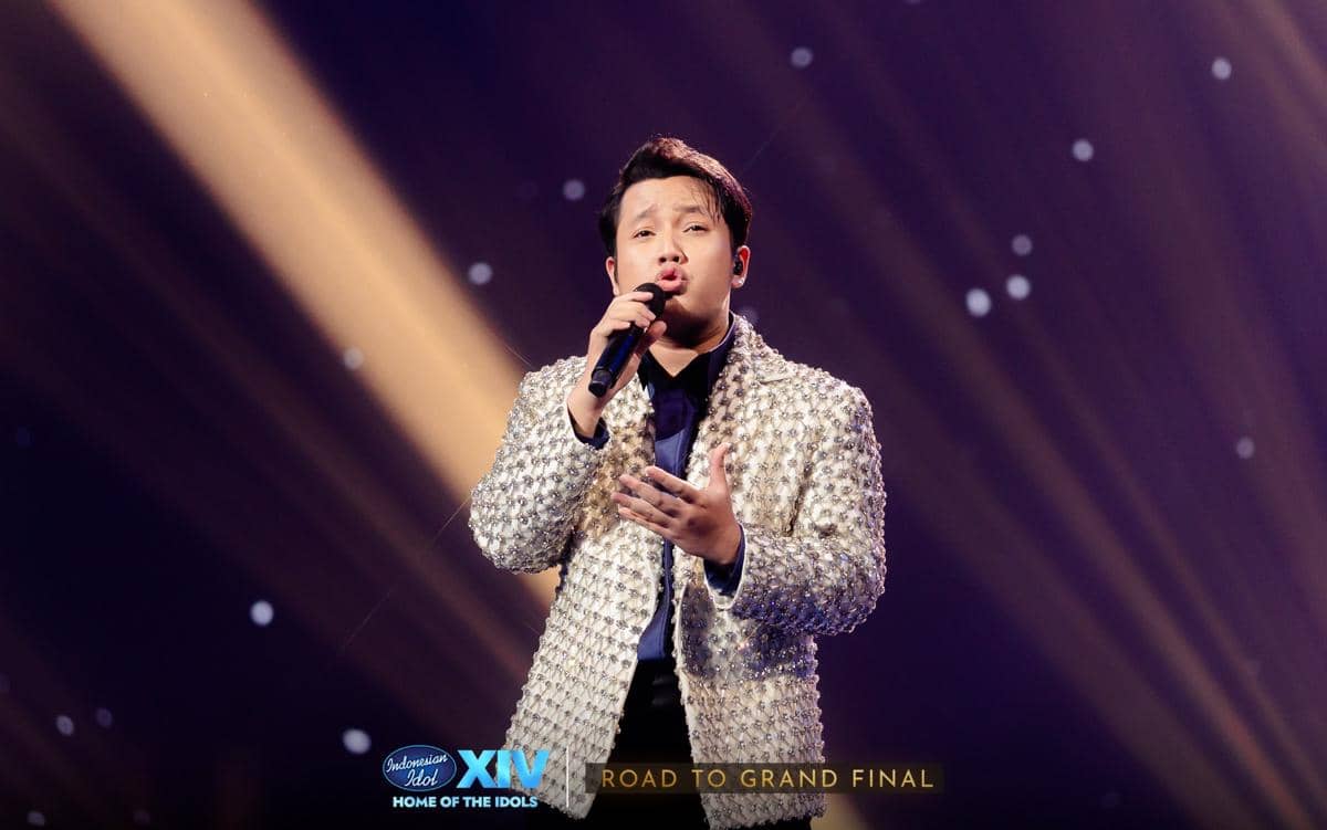 Seorang pria mengenakan jas berkilau sedang bernyanyi di atas panggung dengan pencahayaan dramatis dalam acara Indonesian Idol XIV.