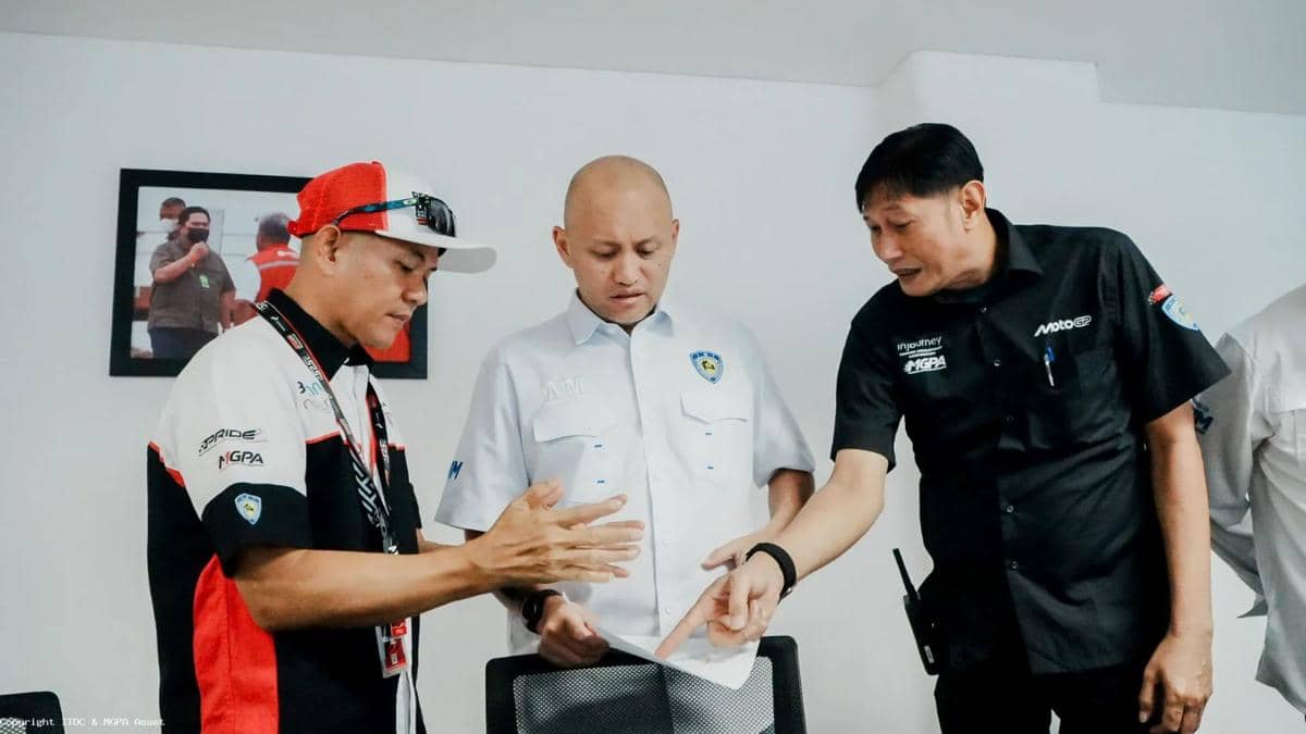Tiga pria berdiskusi sambil melihat dokumen di sebuah ruangan, mengenakan seragam bertema motorsport dengan logo MGPA dan MotoGP.