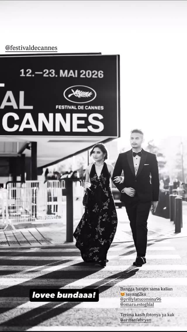 Dua orang berpakaian formal berjalan beriringan di area Festival de Cannes 2026 dengan latar papan besar bertuliskan tanggal acara.