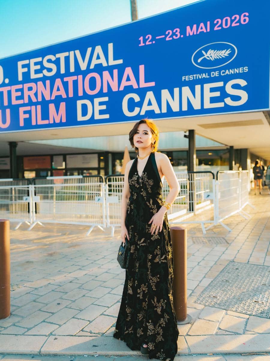 Seorang wanita mengenakan gaun hitam bermotif emas berdiri di depan papan Festival de Cannes 2026 dengan pencahayaan sore hari.