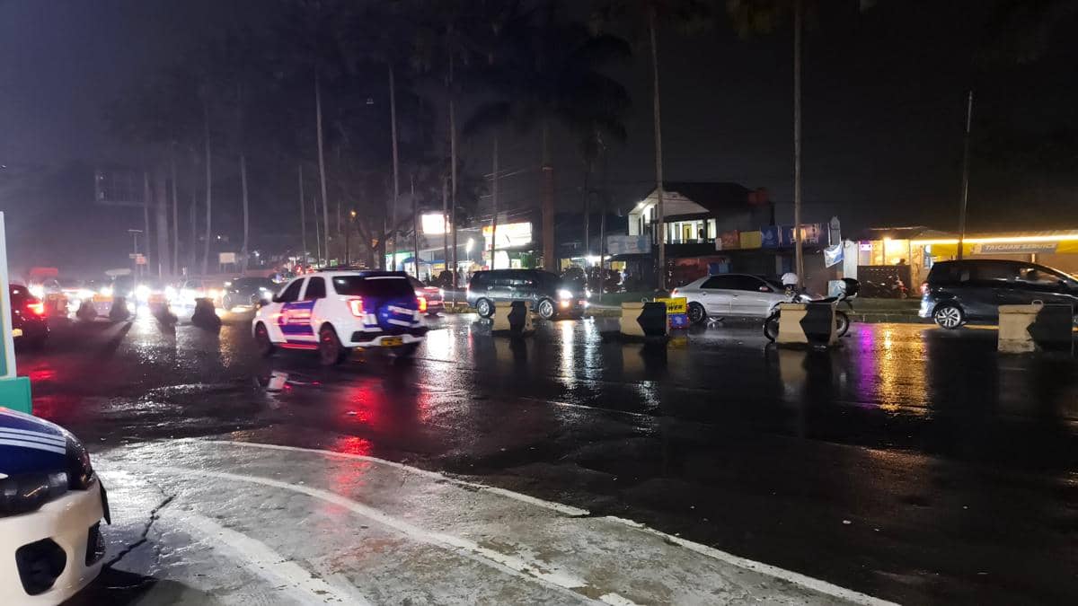 Lalu lintas malam hari di simpang Gadog, Puncak Bogor, dengan kendaraan melintas di jalan basah setelah hujan dan lampu kendaraan memantul di aspal.