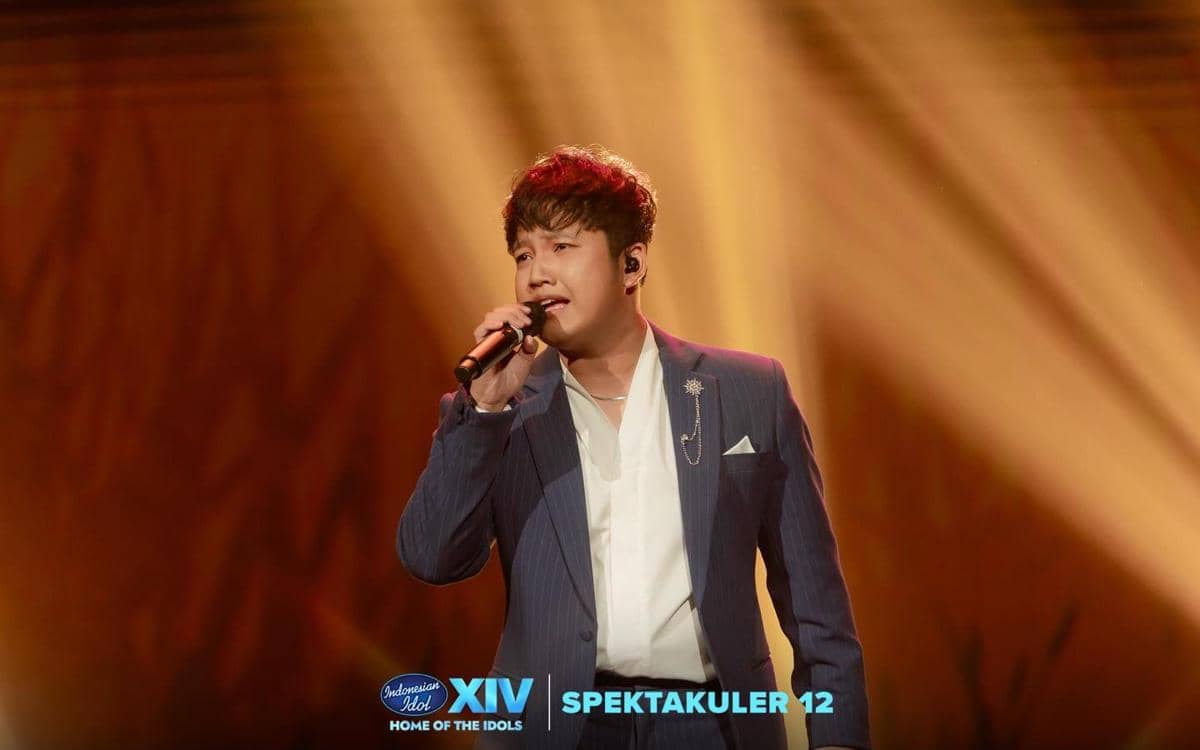 Seorang pria mengenakan setelan jas biru bernyanyi di atas panggung dengan pencahayaan kuning keemasan dalam acara Indonesian Idol XIV Spektakuler 12.