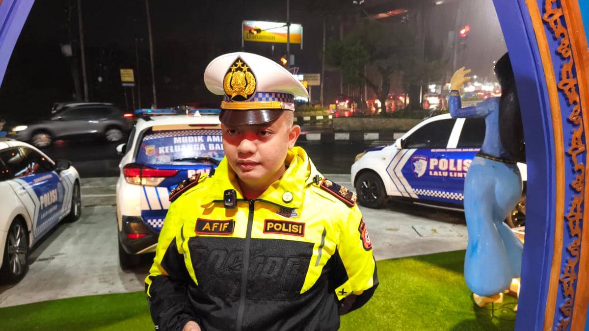 Seorang petugas polisi lalu lintas mengenakan seragam kuning dengan topi dinas berdiri di area jalan malam hari dengan mobil patroli di belakangnya.