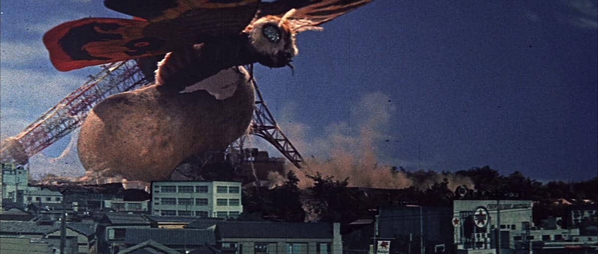 Mothra, makhluk raksasa berbentuk ngengat, melayang di atas kota dengan latar bangunan dan menara yang runtuh akibat kehancuran.