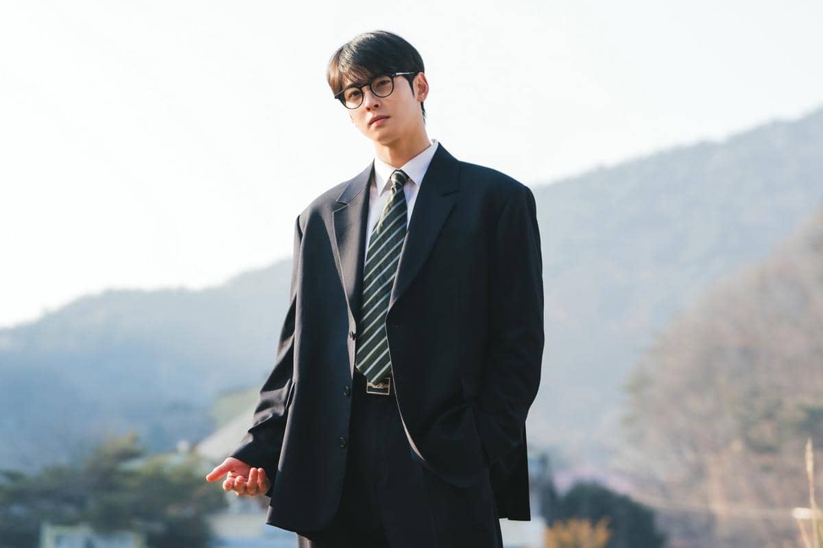 Seorang pria mengenakan setelan jas hitam dan dasi bergaris berdiri di luar ruangan dengan latar pegunungan dalam adegan drama Korea The WONDERfools.