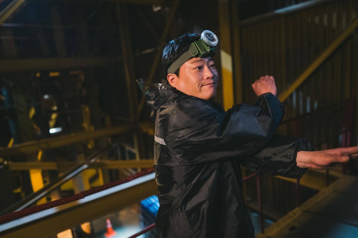 Seorang pria mengenakan jaket hitam dan lampu kepala sedang bersiap melakukan gerakan di area industri dengan pencahayaan redup.