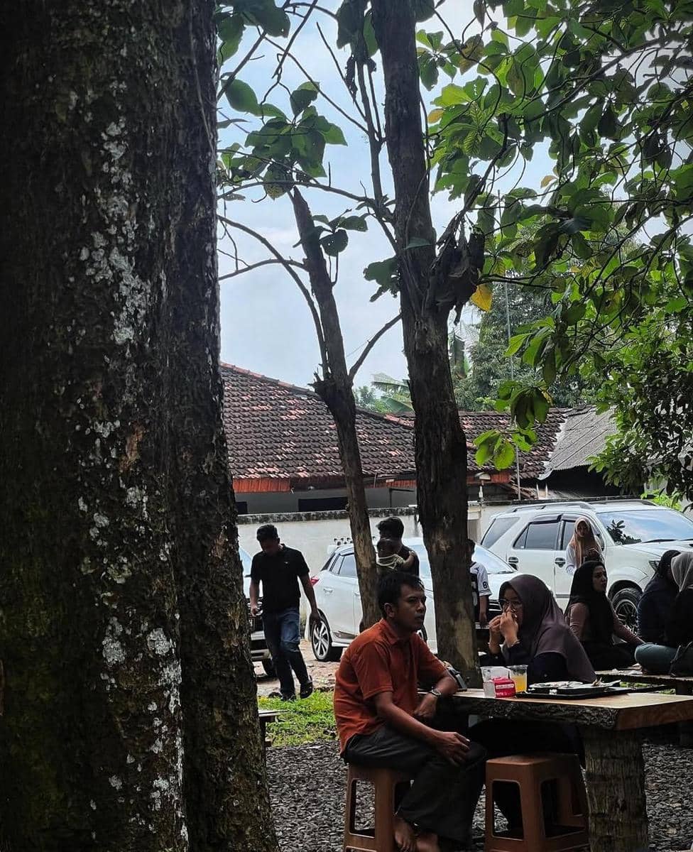 Beberapa orang menikmati hidangan di area outdoor Pawon Numani Kota Metro dengan suasana rindang di bawah pepohonan besar.