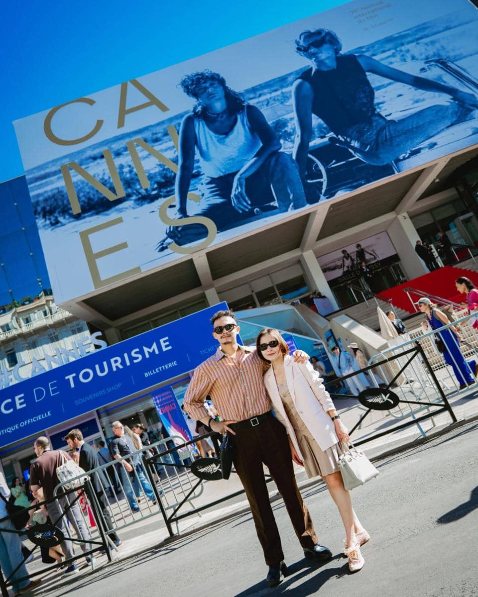 Dua orang berpose di depan gedung utama Cannes Film Festival dengan latar spanduk besar bertuliskan CANNES di bawah langit cerah.