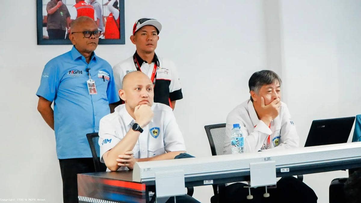 Beberapa pria mengenakan seragam resmi duduk dan berdiri di ruang rapat sambil memperhatikan layar laptop di depan mereka.