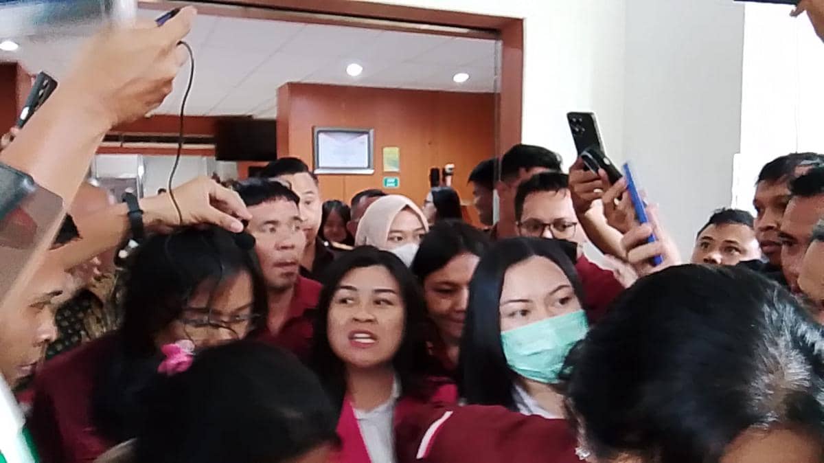 Kerumunan orang di dalam gedung Kejati Sulut, beberapa mengenakan pakaian merah dan masker, dikelilingi wartawan yang memegang ponsel dan mikrofon.