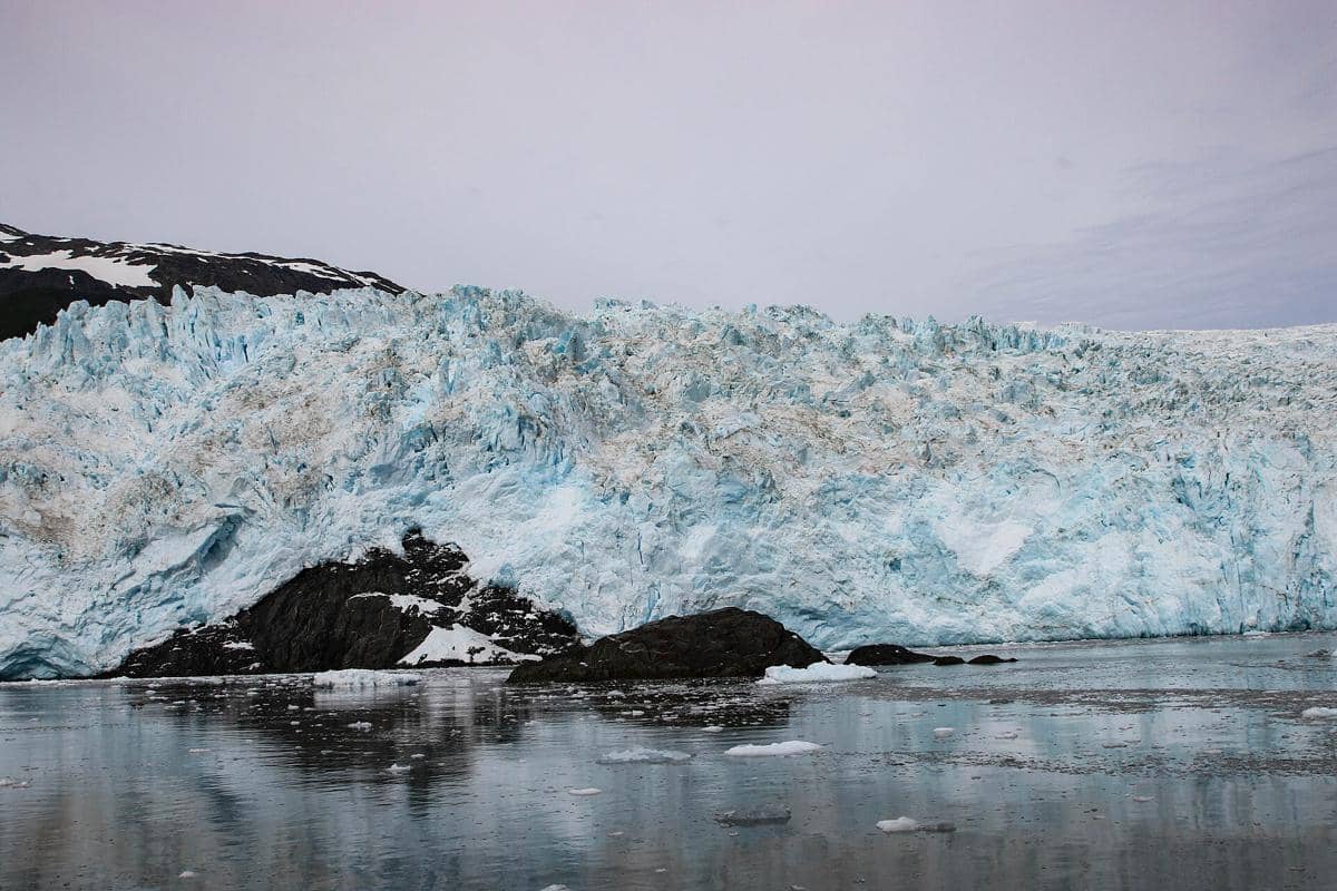 Gletser besar berwarna biru muda di Kenai Fjords dengan bongkahan es mengapung di permukaan air dan langit mendung di latar belakang.