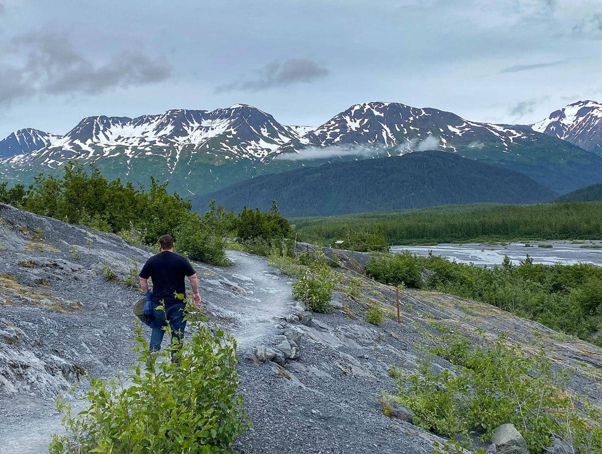 Seorang pria berjalan di jalur pendakian berbatu dengan latar pegunungan bersalju di Kenai Fjords, Alaska, di bawah langit berawan.