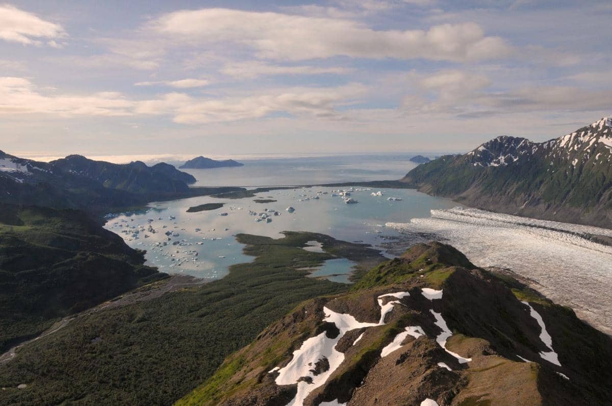 Pemandangan udara Kenai Fjords dengan gletser besar, pegunungan bersalju, dan potongan es yang mengapung di laut biru jernih.